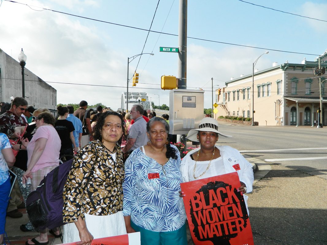 Three women holding signs standing in front of a crowd.