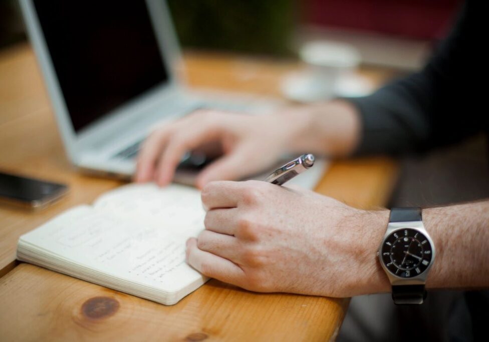 A person is writing on paper with a laptop.